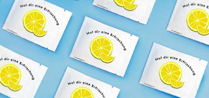 Erfrischungstücher mit Logo liegen auf einem blauen Tisch. Auf der Verpackung der bedruckten Erfrischungstücher sind Zitronen abgebildet.
